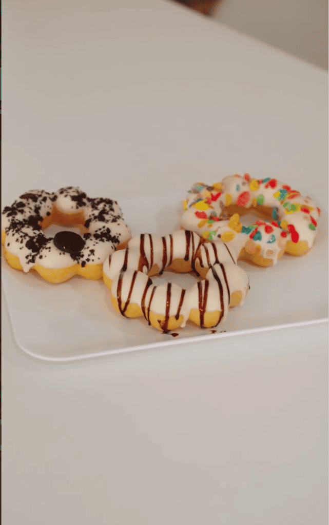 Four decorated mini donuts topped with white icing, oreo crumbles, chocolate drizzle, and colorful cereal sprinkles are arranged on a white plate.