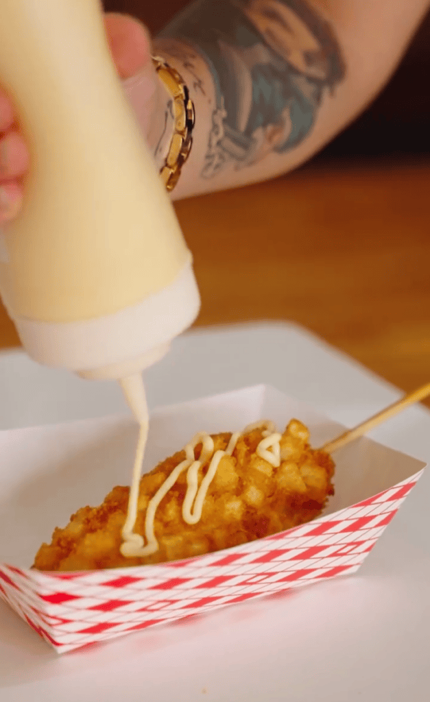 A person drizzles creamy sauce from a squeeze bottle onto a piece of fried food served in a red and white checkered paper tray.