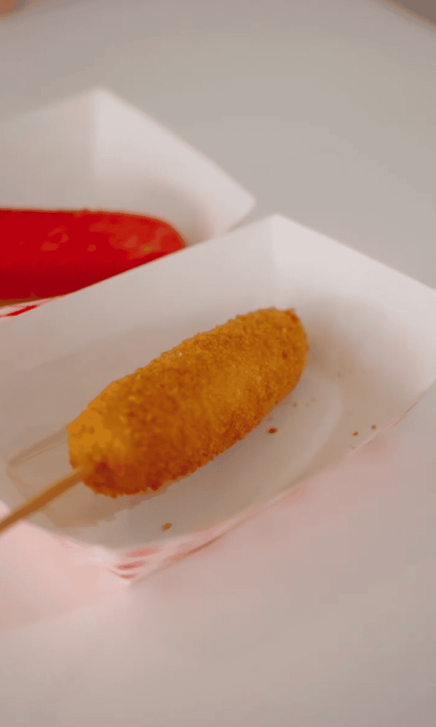 A close-up of a breaded corn dog on a stick served in a paper tray with a side of ketchup.