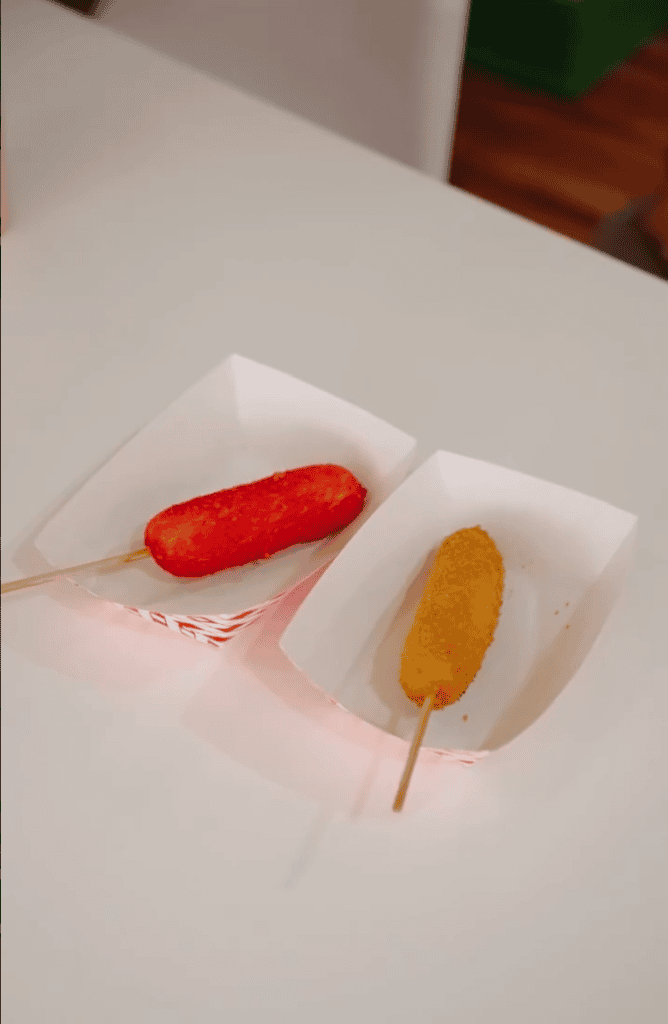 Two corn dogs on sticks, one red-coated and one golden-breaded, sit on paper trays on a white table.