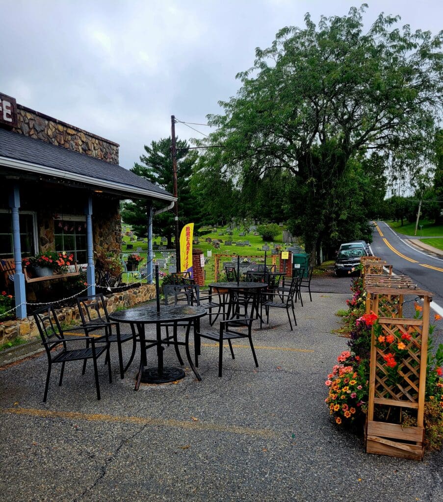 A peaceful outdoor café patio with empty seating, flowering planters, and a large tree, suggesting a quiet moment at a small restaurant or shop.