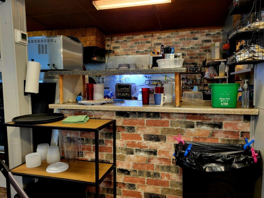 A cluttered home workshop or garage with exposed brick walls, work tables, shelving units, and various tools and equipment organized on surfaces.