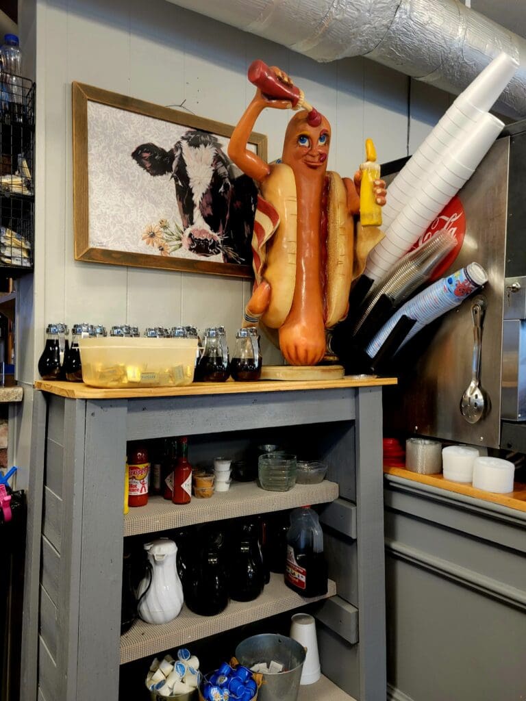 A gray kitchen storage cabinet displaying a large hot dog sculpture on top alongside wine bottles, with shelves below containing various kitchen items and food products.