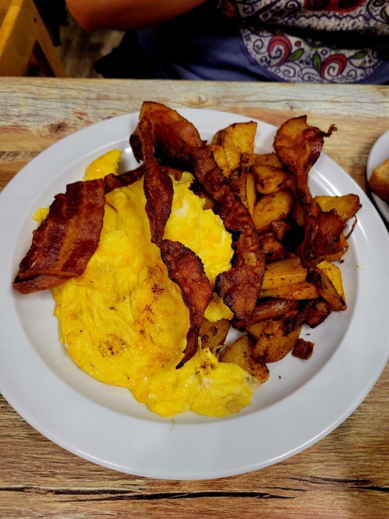 A breakfast plate featuring scrambled eggs, crispy bacon, and roasted potatoes served on a white plate on a wooden table.