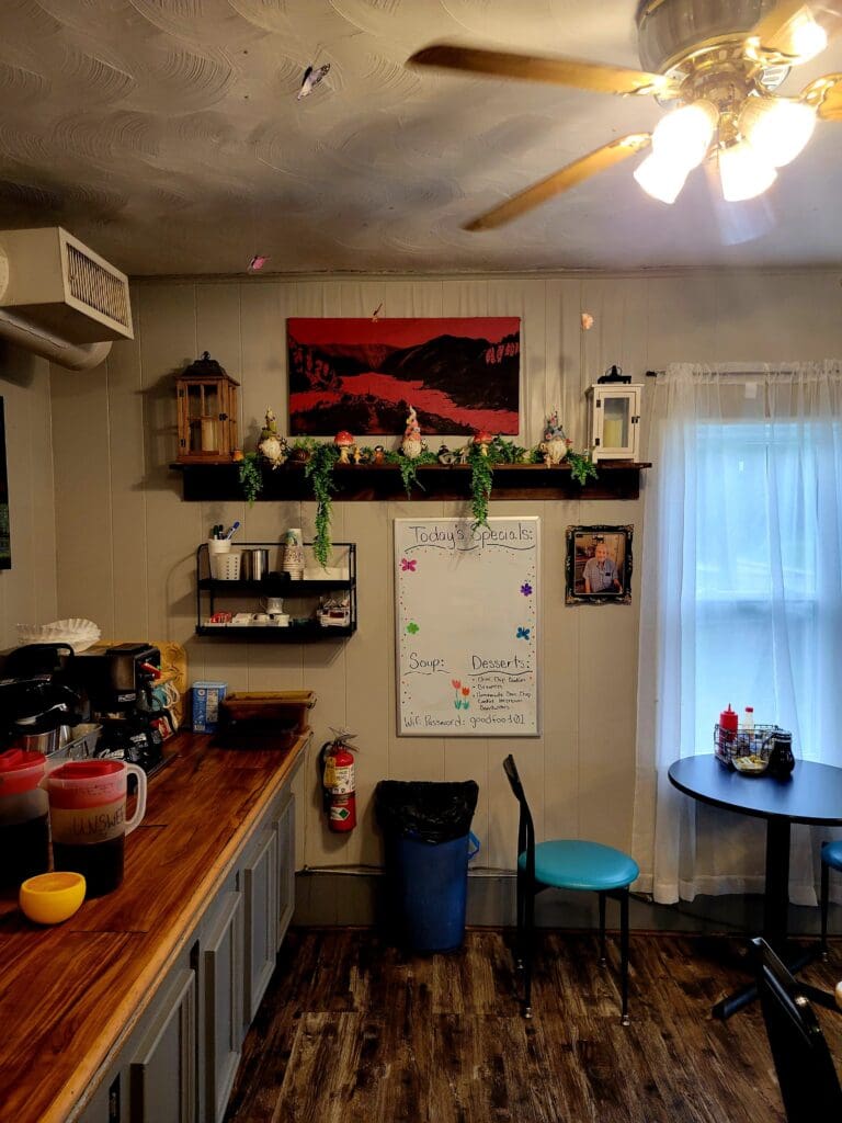 A cozy, modestly decorated kitchen interior featuring wooden countertops, a refrigerator, ceiling fan, and various decorative items on shelves.