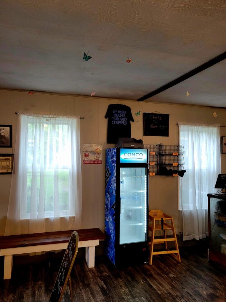 A quiet restaurant or café interior with wooden furniture, a beverage cooler, natural light from sheer curtains, and rustic décor on the walls.