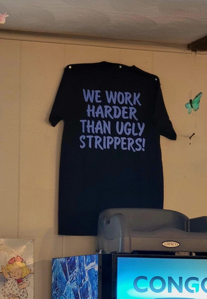 A dark navy t-shirt with blue text reading 'WE WORK HARDER THAN UGLY STRIPPERS!' is displayed on a wall in an indoor workshop or office space.
