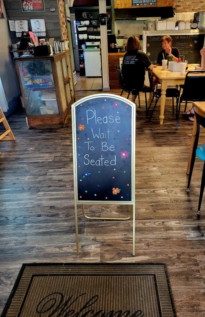 A chalkboard sandwich board sign with handwritten text sits in the entrance of a wooden-floored venue or restaurant, with people visible in the background.