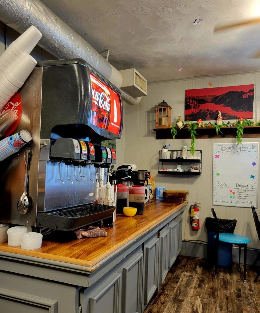 A cozy indoor coffee shop workspace featuring an espresso machine, wooden counter, shelving with supplies, and neon signage in a compact café setting.