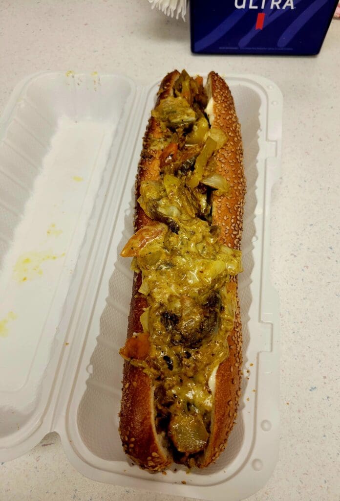 A hot dog topped with mustard and relish sits in a white foam container on a white surface.