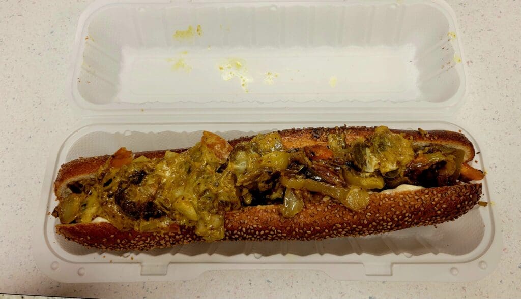 A grilled hot dog topped with yellow mustard and relish served in a clear disposable container.