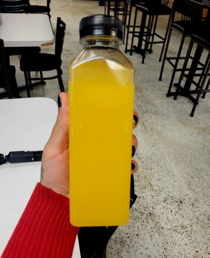 A person in a red sleeve holds a bottle of bright yellow beverage in a casual restaurant or cafΓ© setting.