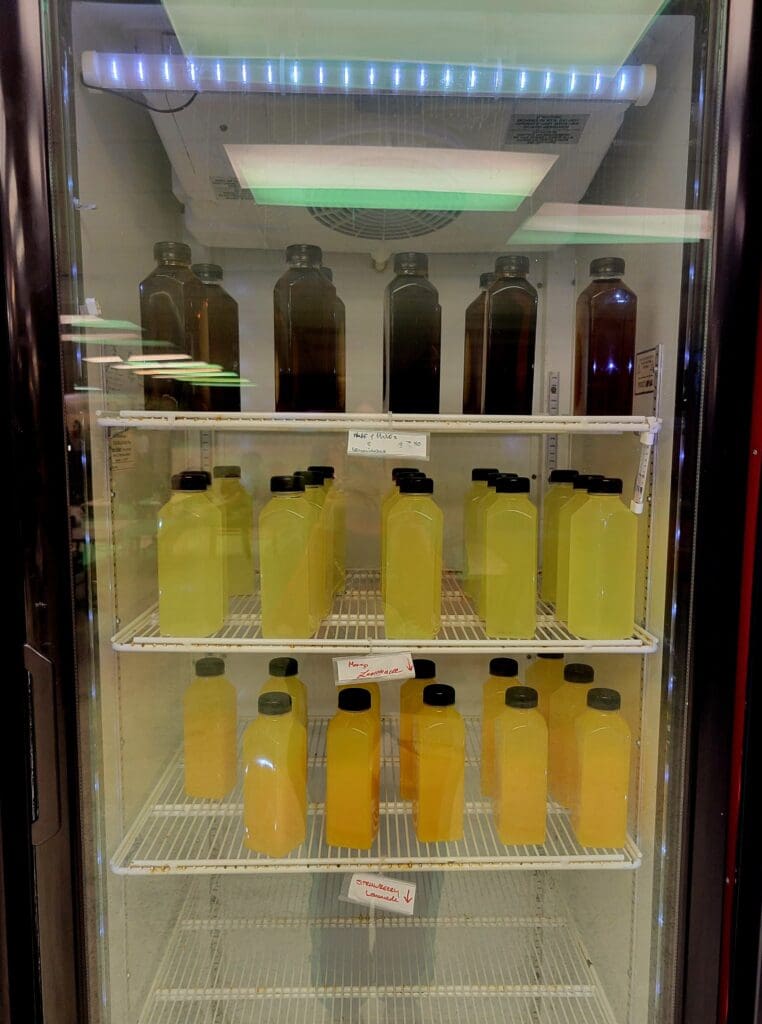 A glass-front refrigerator display case stocked with multiple shelves of bottled beverages in yellow and amber-colored liquids, illuminated by blue LED lighting at the top.