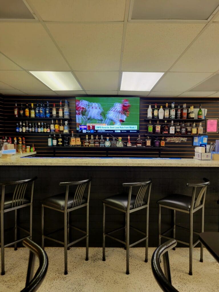 An empty bar counter with dark seating, well-stocked liquor shelves, and a mounted television screen in an indoor venue setting.