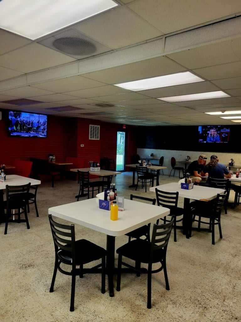 A casual indoor community or event space with multiple dining tables, chairs, and a bar area, featuring neutral tones and overhead lighting.