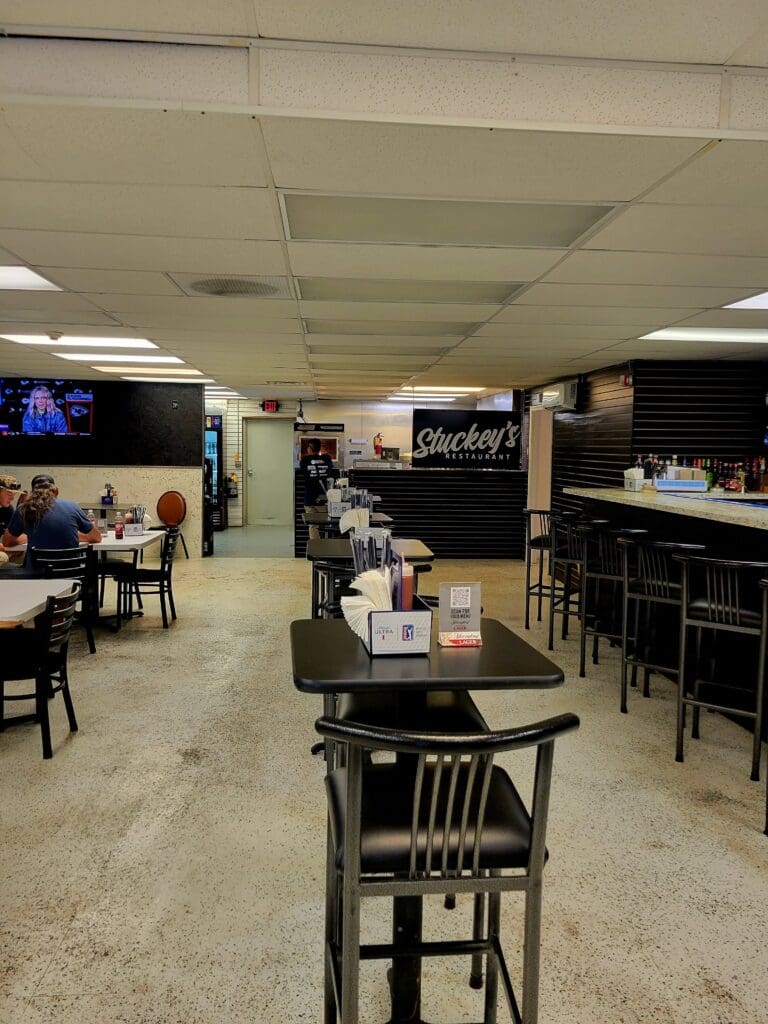 An empty indoor workshop or community space with tables, black chairs, shelving units, and a kitchen area beneath industrial ceiling lights.