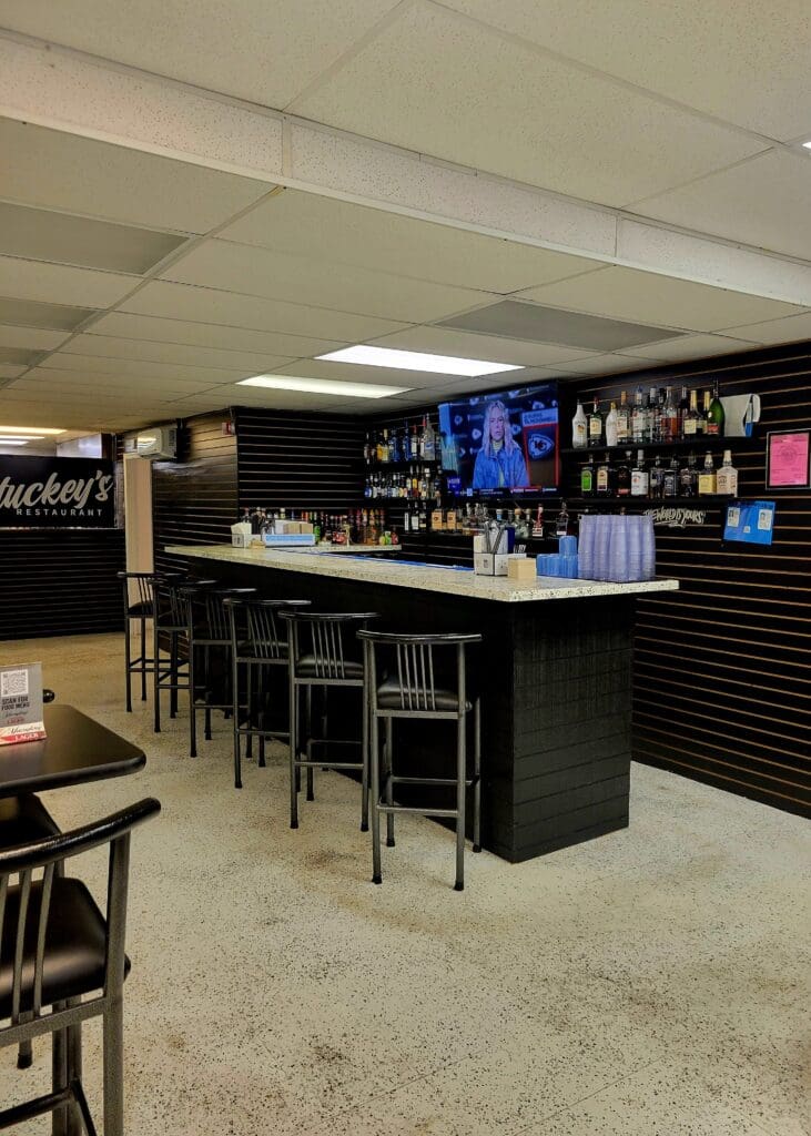 An empty modern bar venue with a sleek black counter, bar seating, and well-stocked shelving under ambient lighting.