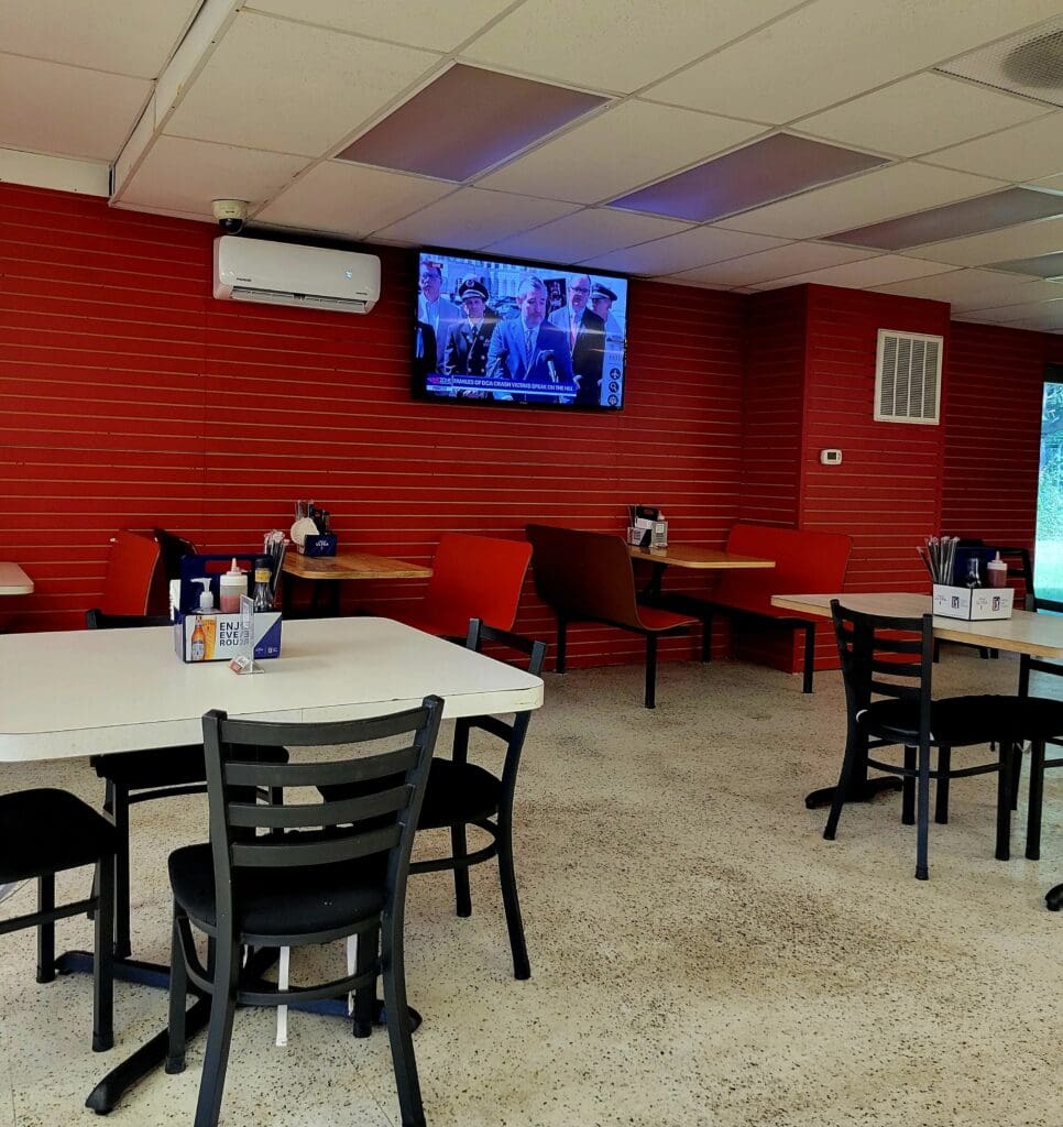 A modern meeting or training room with bright red brick walls, multiple white tables with black chairs, and a wall-mounted television display.