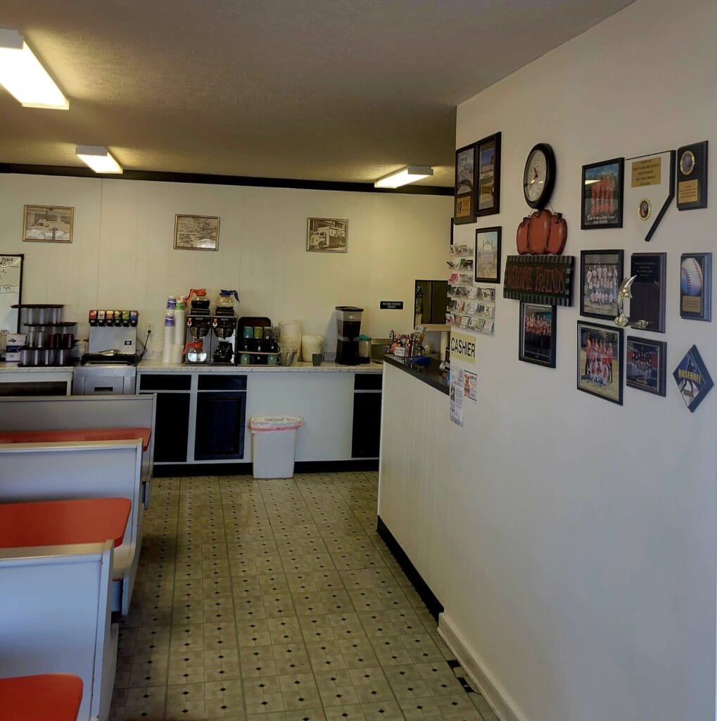 An empty small-town diner interior with red booth seating, a coffee and drink station, checkered tile floor, and a wall decorated with sports plaques, photos, and a clock near the cashier counter.