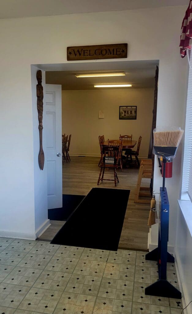 A doorway with a wooden 'Welcome' sign leads into a dining or meeting room with wooden tables, chairs, and a framed picture on the wall.
