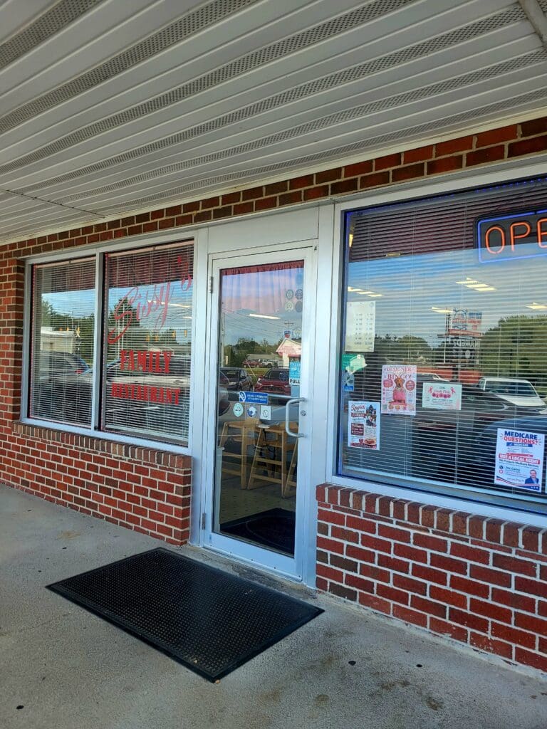 The exterior entrance of a small family restaurant with a red brick facade, glass door, and a neon 'open' sign in the window.
