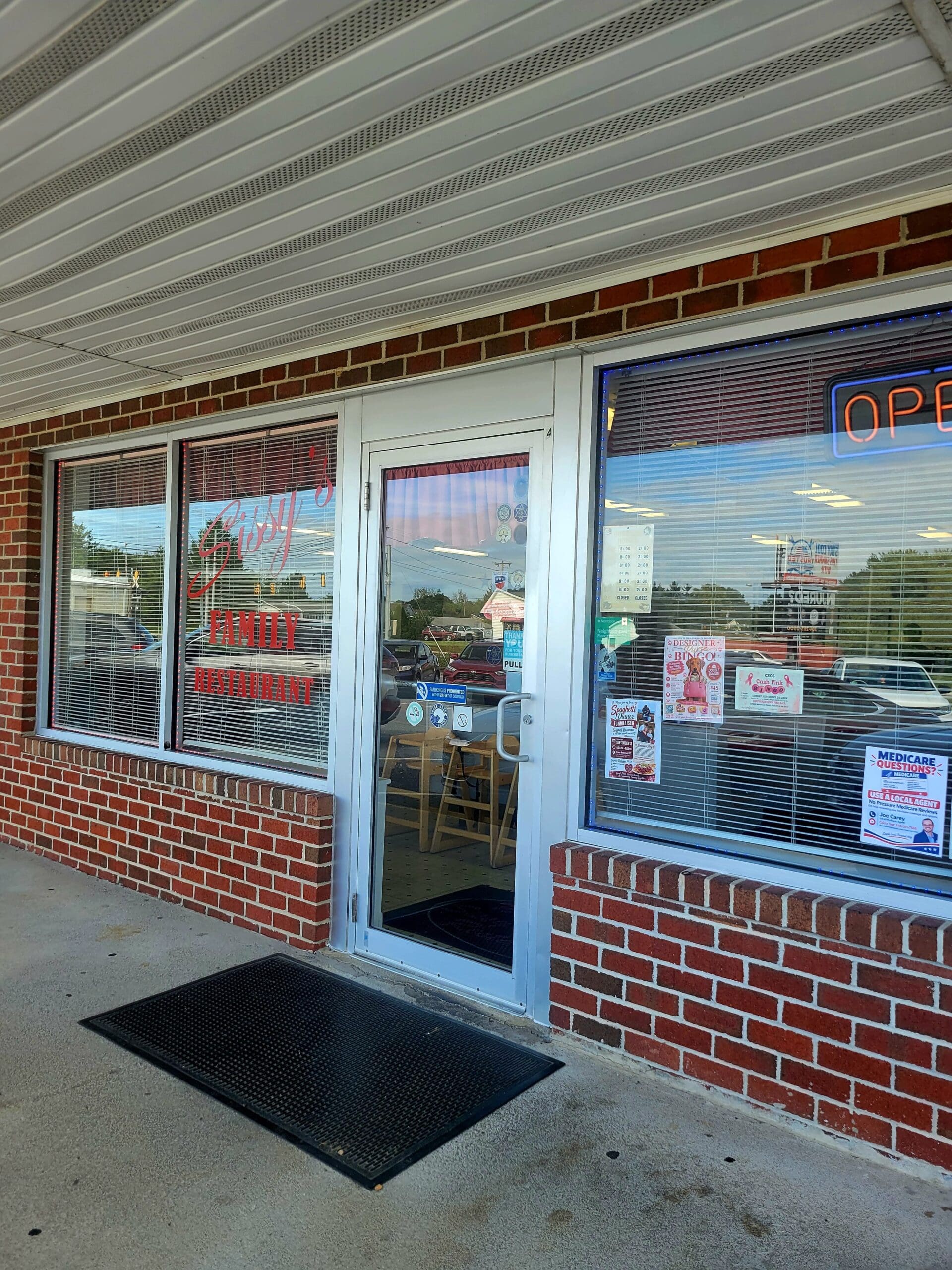 The exterior entrance of a small family restaurant with a red brick facade, glass door, and a neon 'open' sign in the window.