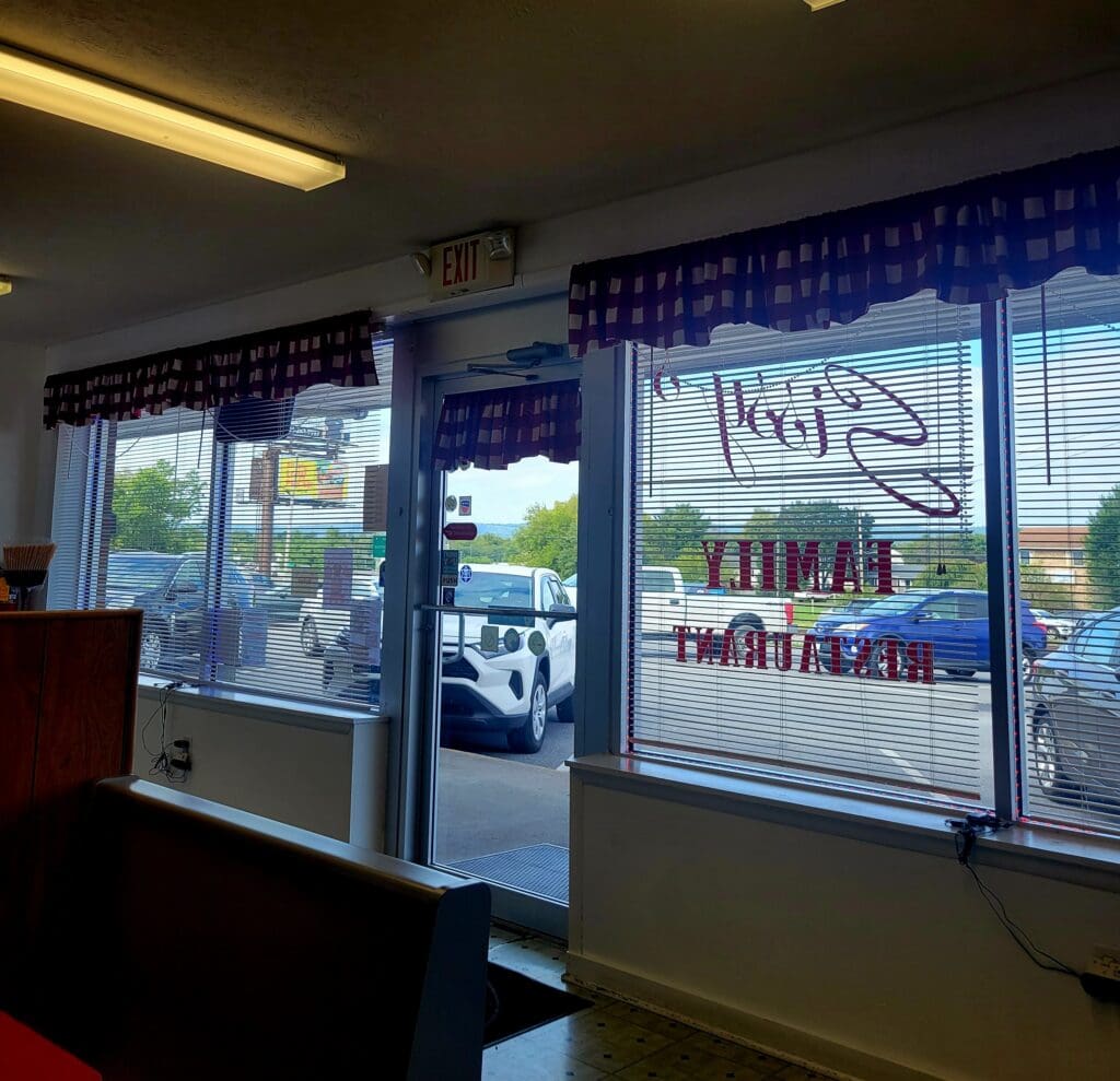Interior view of a family restaurant looking out through window blinds and a glass door toward a parking lot with several cars.