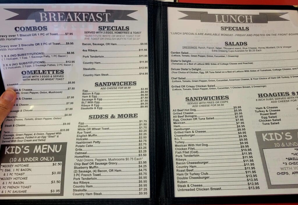 A photo of an open breakfast and lunch menu at a diner, with a hand holding the page open, listing combos, omelettes, specials, sandwiches, salads, and hoagies with prices.