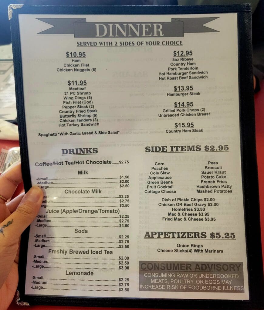 A person holds open a laminated dinner menu listing entrees, side items, drinks, and appetizers with prices.