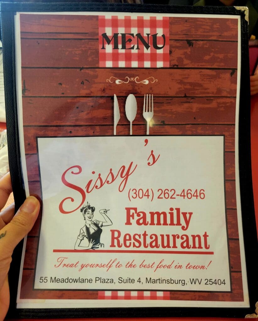 A person holds open a menu cover for Sissy's Family Restaurant featuring a wood-plank design and red gingham accents.