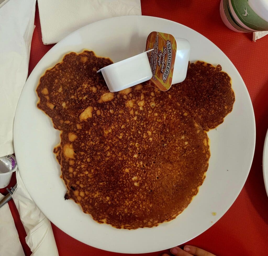 An overhead shot of a Mickey Mouse-shaped pancake on a white plate with a small Smucker's syrup container, set on a red table.