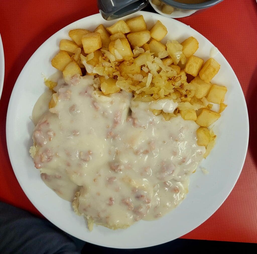 An overhead shot of a diner plate with biscuits smothered in sausage gravy alongside crispy home fries and sauerkraut, set on a red table.