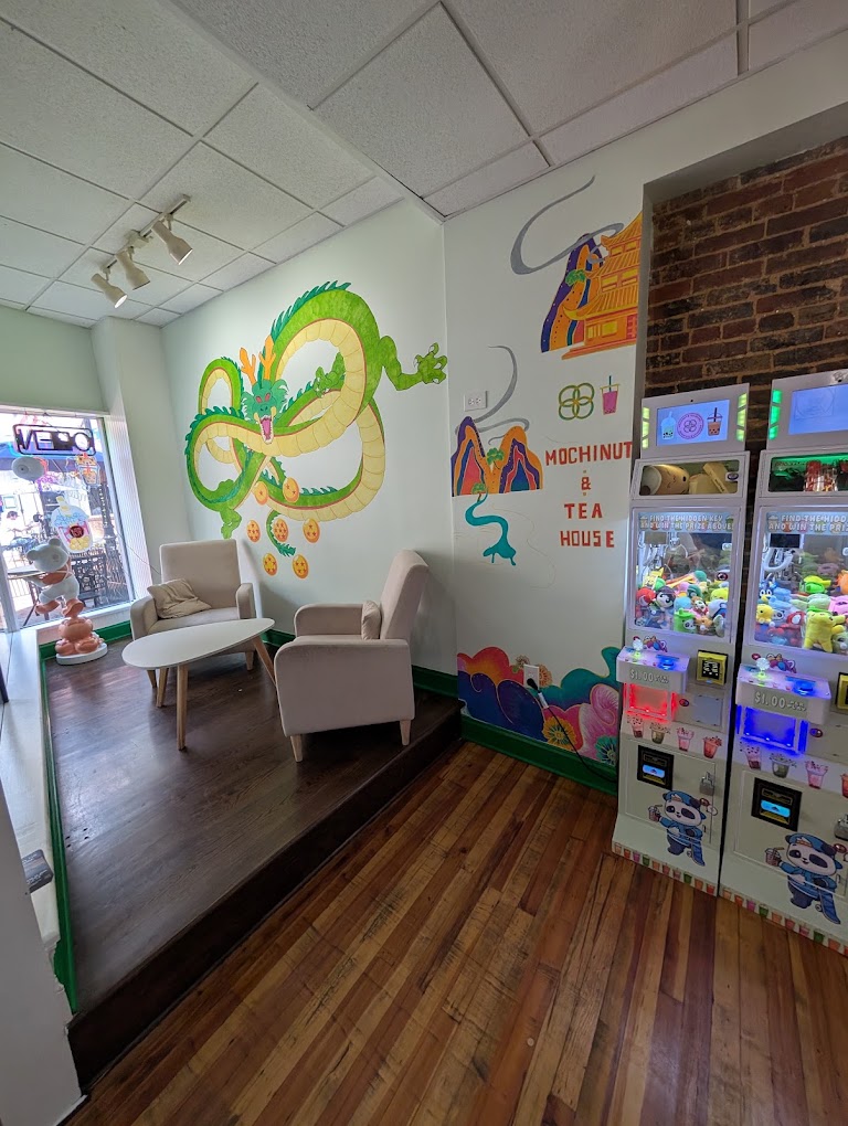 An interior seating area of a Mochinut and Tea House featuring a colorful dragon mural, cozy armchairs, and arcade claw machines.