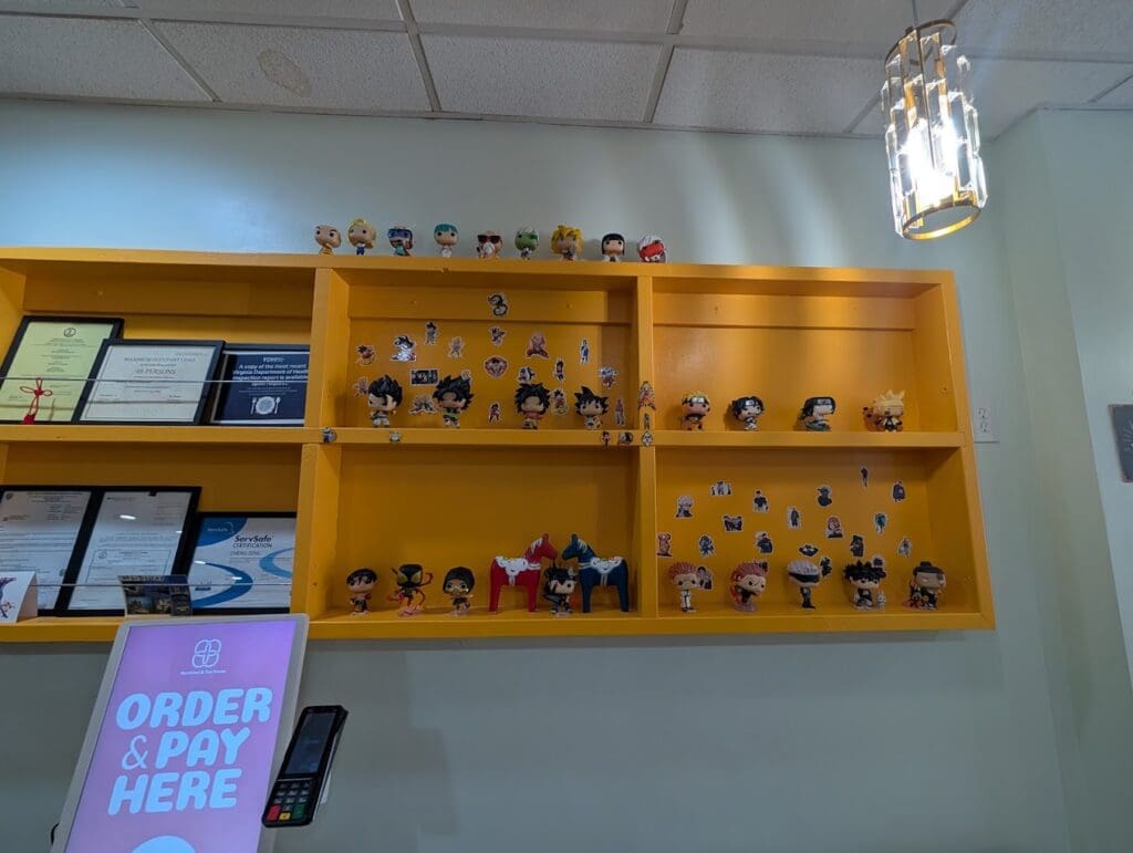 A mustard-yellow display shelf in a restaurant holds anime-themed Funko Pop figures and stickers above certifications, with an 'Order & Pay Here' sign visible below.