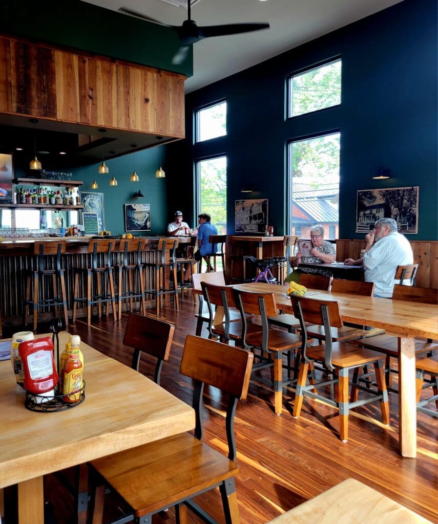 A cozy restaurant interior with wooden tables and bar stools, dark teal walls, large windows, and a few patrons dining.