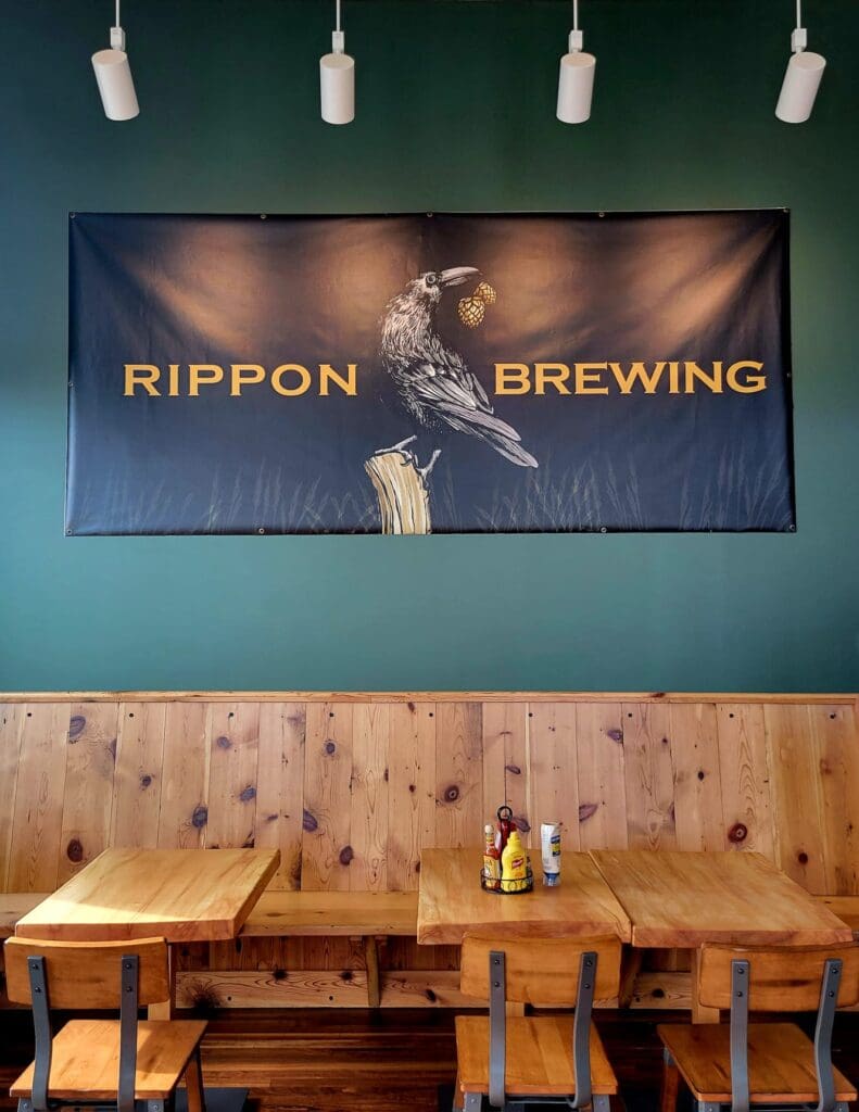 An empty restaurant or brewery interior with wooden tables and chairs beneath a large 'Rippon Brewing' banner featuring a crow illustration on a teal wall.