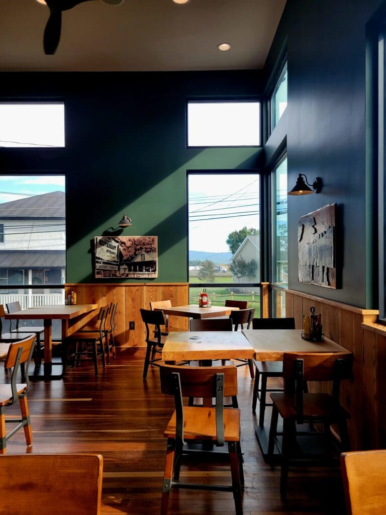 An empty restaurant dining area with wooden tables and chairs, dark green walls, large windows letting in daylight, and framed artwork.