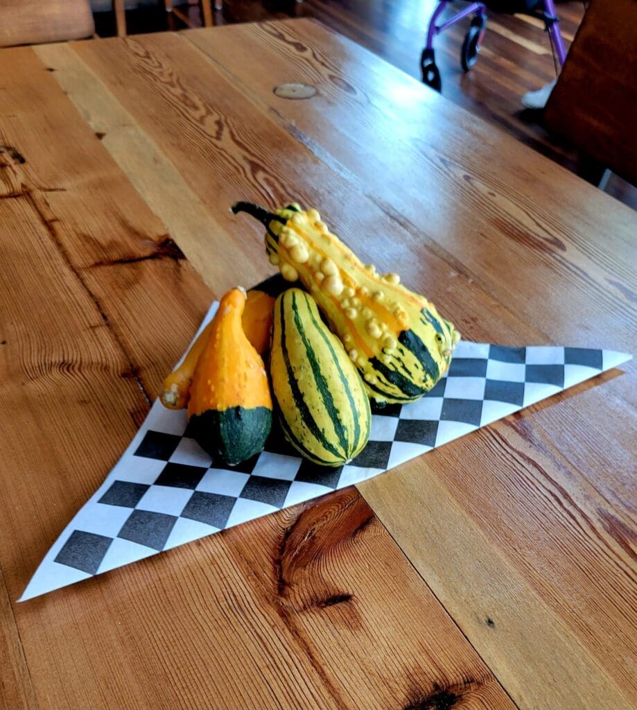 A trio of decorative autumn gourds sits on a black-and-white checkered paper atop a wooden table, with a walker visible in the background.