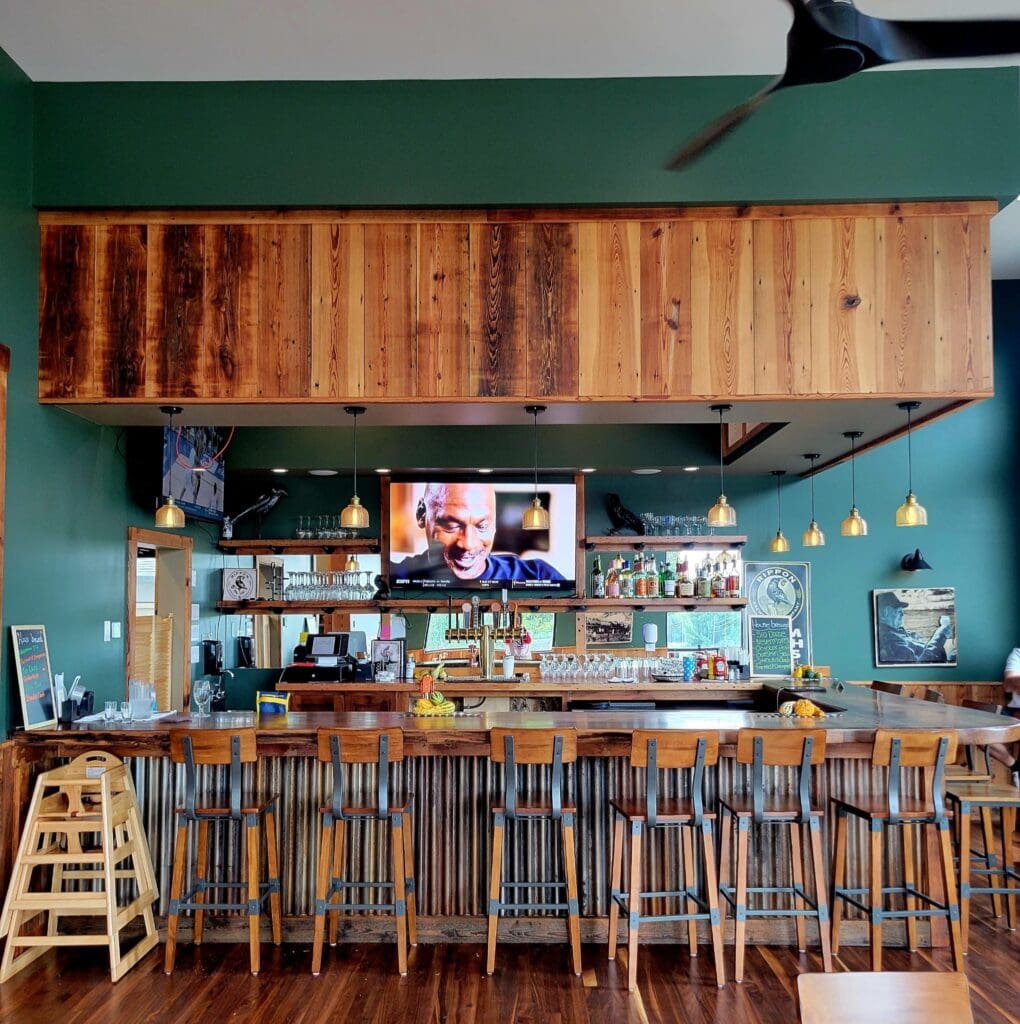 An empty wooden bar with stools, hanging pendant lights, and a TV mounted above the liquor shelves in a teal-walled restaurant or pub.