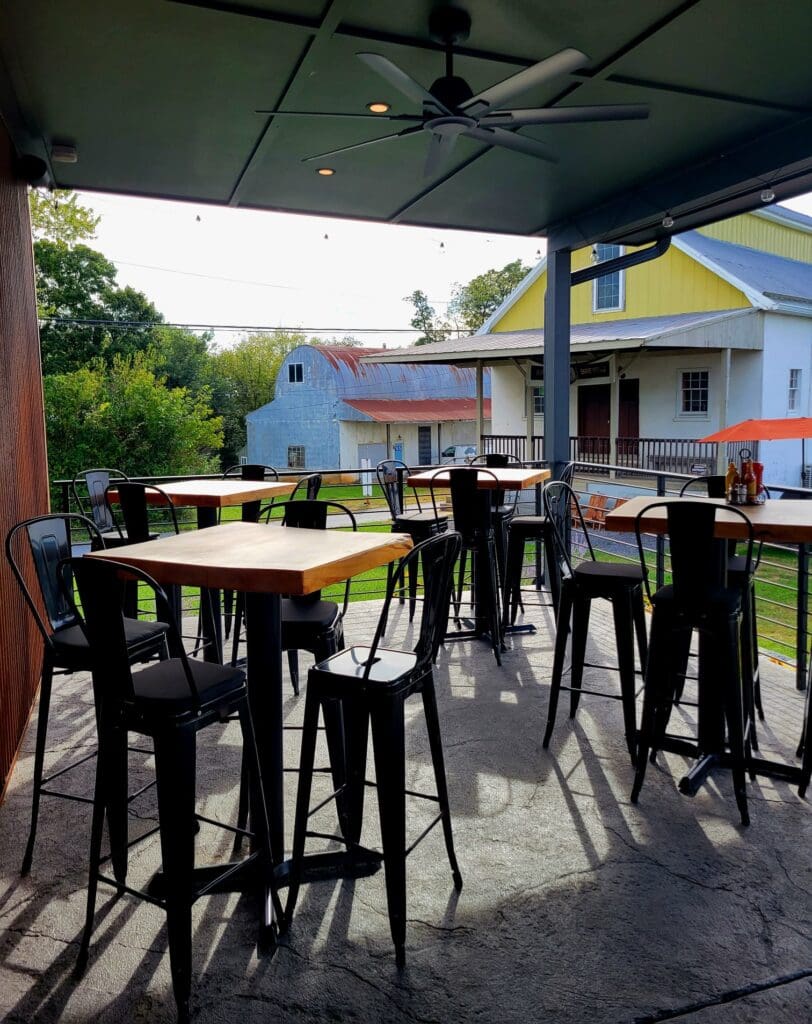 An empty covered outdoor patio with high-top wooden tables and black metal bar stools overlooking a rural scene with a barn.