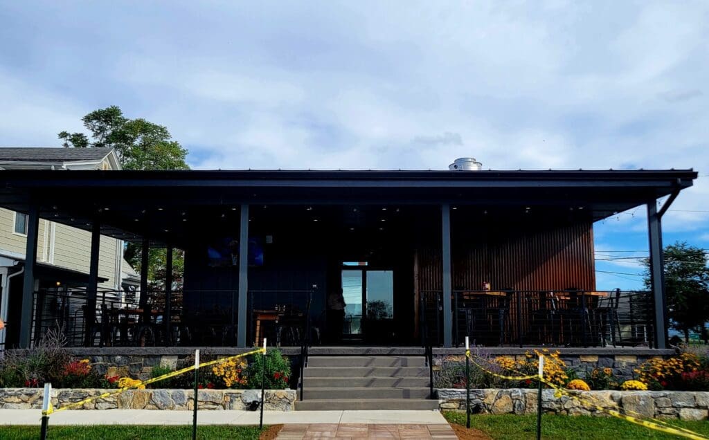 A covered outdoor patio dining area at a restaurant with black metal framing, stone retaining wall, and flower beds under a cloudy sky.