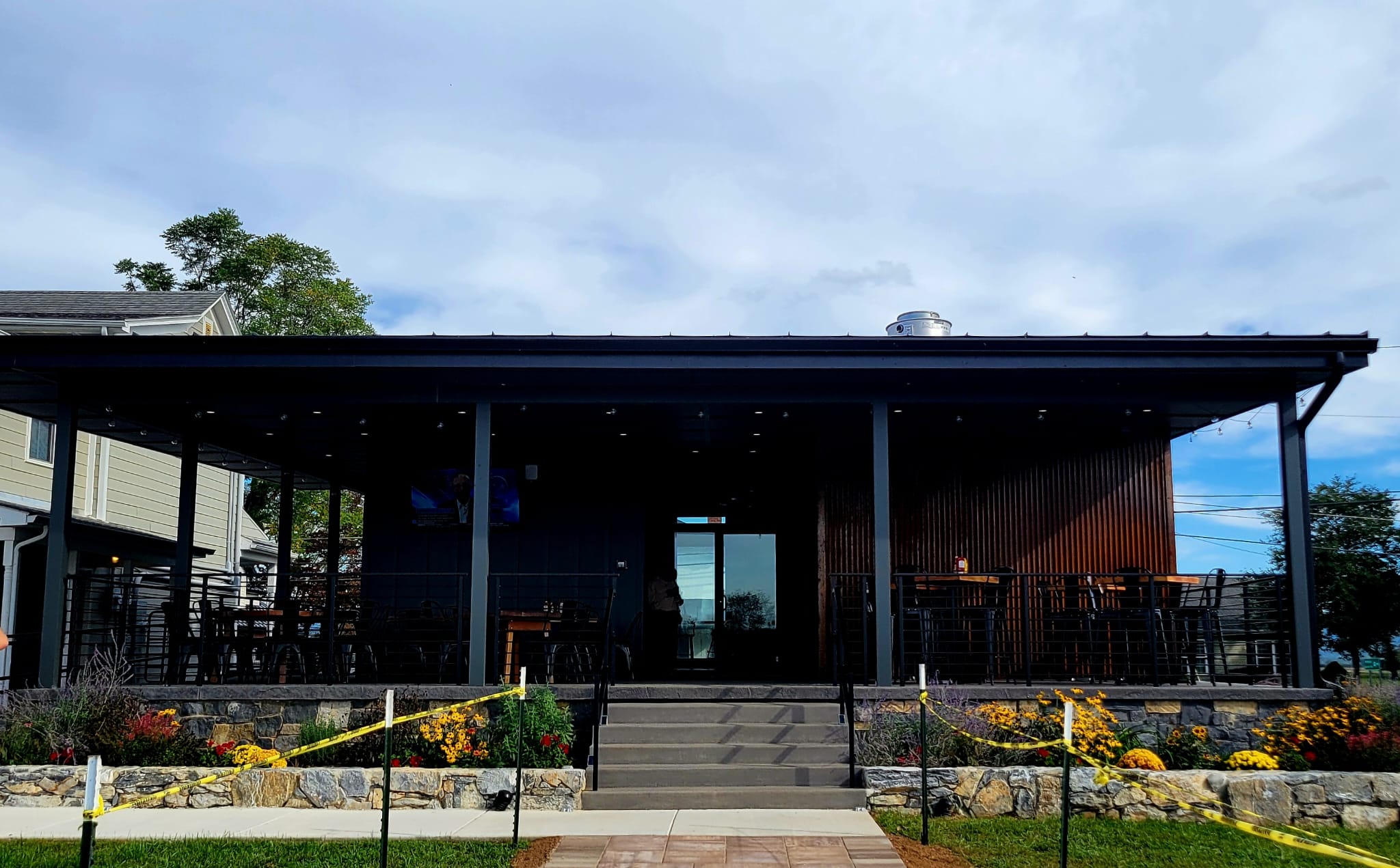 A covered outdoor patio dining area at a restaurant with black metal framing, stone retaining wall, and flower beds under a cloudy sky.