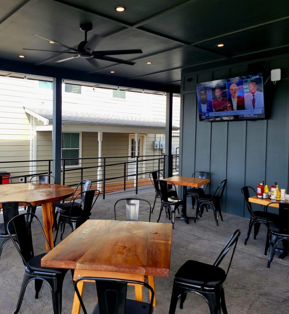 An empty outdoor restaurant patio with wooden tables, black metal chairs, a ceiling fan, and a wall-mounted TV showing a news broadcast.