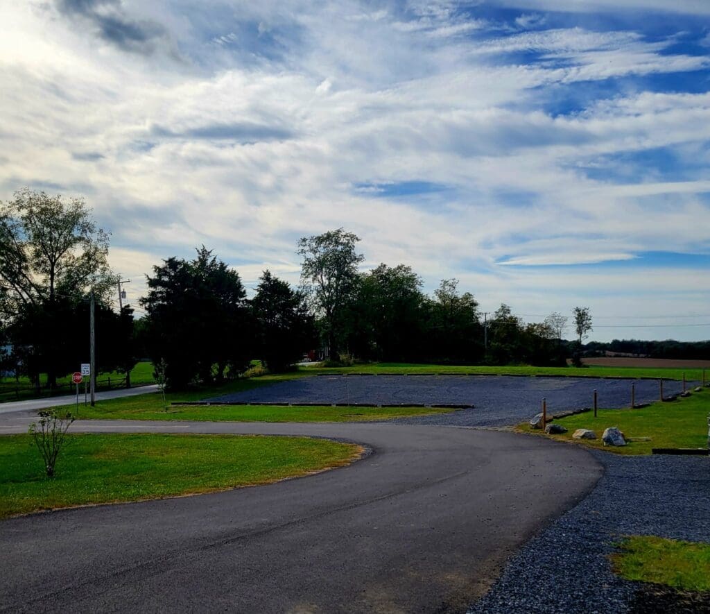 A paved driveway curves through a grassy rural property leading to a gravel parking area under a partly cloudy sky.