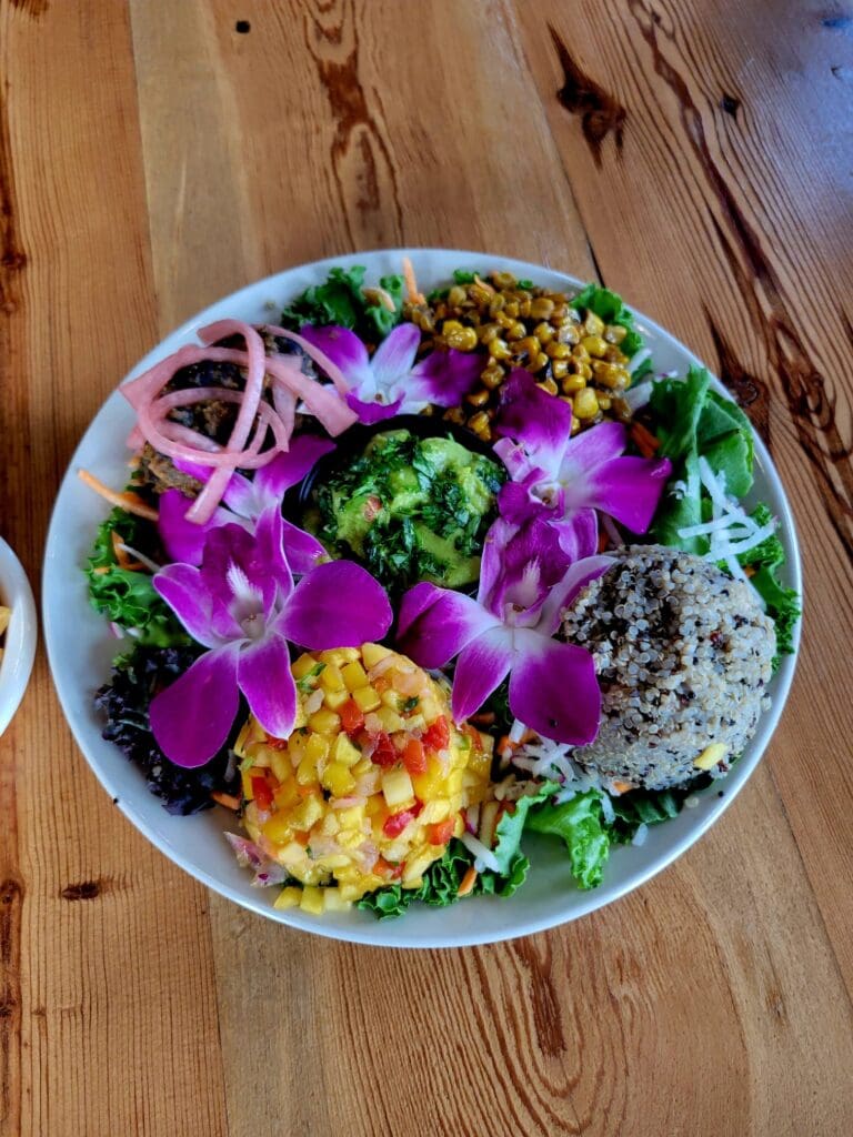 An overhead shot of a colorful gourmet salad bowl garnished with purple orchids, mango salsa, guacamole, quinoa, pickled onions, and corn on a wooden table.