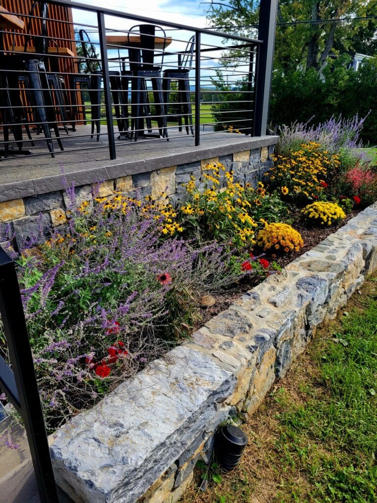 A landscaped stone retaining wall with colorful flower beds sits below a deck with bar stools and railing, overlooking a grassy lawn and trees.