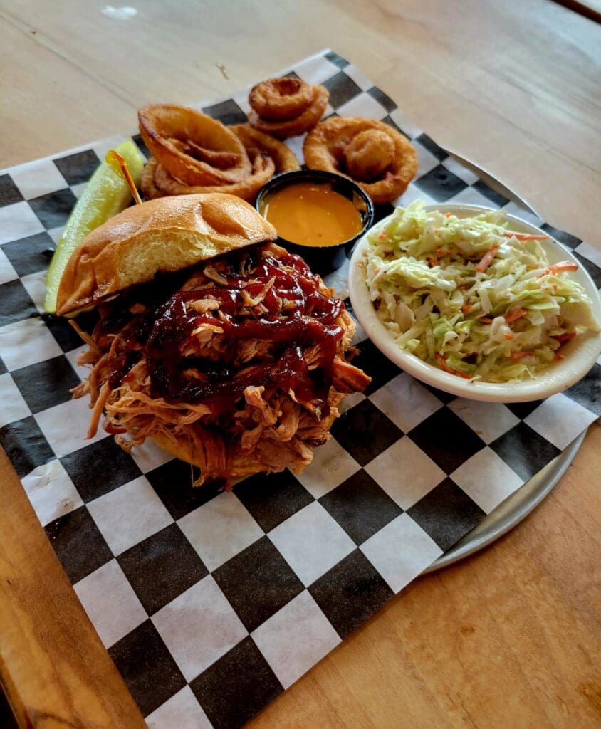 An overhead shot of a barbecue pulled pork sandwich with onion rings, coleslaw, a pickle spear, and dipping sauce served on a checkered paper-lined tray.