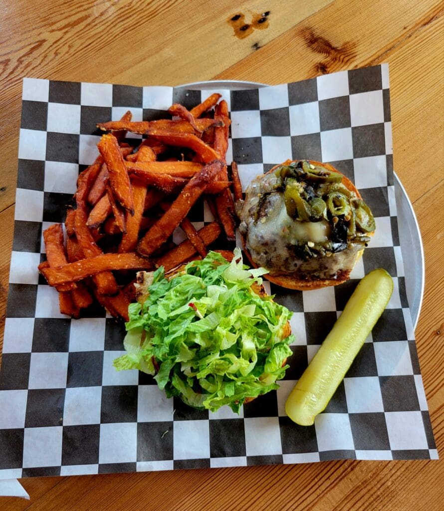 An overhead shot of a cheeseburger topped with jalapenos and lettuce, served with sweet potato fries and a pickle spear on checkered paper.