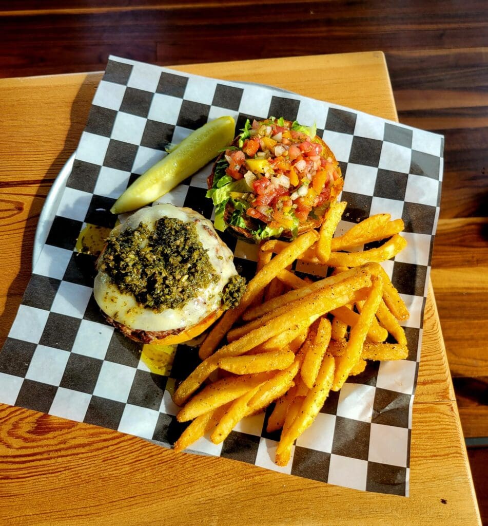 An overhead shot of a pesto cheeseburger with a pickle spear and seasoned french fries served on checkered paper.