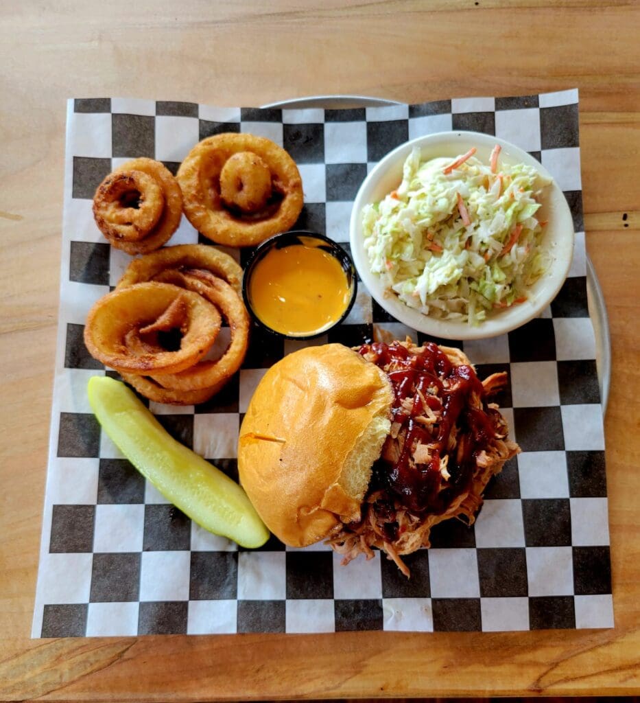 An overhead shot of a pulled pork sandwich with barbecue sauce, served with onion rings, cheese dip, coleslaw, and a pickle spear on a checkered paper-lined tray.